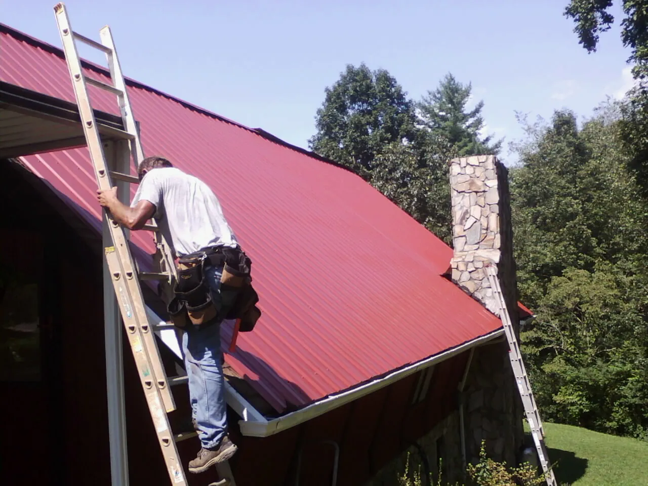 Roofer on a ladder beside a red metal roof and stone chimney — Soffit & Fascia Repair by Roofing Brothers in Wimbledon